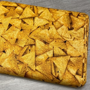 May include: A folded fabric featuring a repeating pattern of golden-brown tortilla chips. The chips are triangular with a textured surface and appear to be seasoned. The fabric is likely for crafting or home decor.