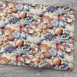 May include: Fabric with a seashell pattern. The design features various seashells, starfish, and coral in shades of orange, brown, blue, and purple, set against a sandy background. The fabric is likely used for crafting or home decor projects.
