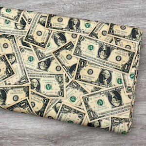 May include: A folded piece of fabric featuring a repeating pattern of United States one-dollar bills. The print displays the portrait of George Washington, the text One Dollar, and the United States of America in black ink on a cream background.