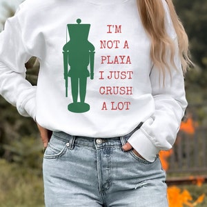 May include: White sweatshirt featuring a green silhouette of a nutcracker and the text "I'm not a playa I just crush a lot" in red.