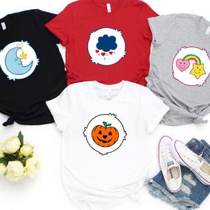 May include: A collection of t-shirts in black, red, white, and gray, each featuring a different cartoon graphic. Designs include a moon and star, a cloud with hearts, a heart and star with a rainbow, and a jack-o'-lantern. White sneakers and denim shorts are also in the image.