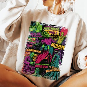 May include: Cream-colored sweatshirt featuring a colorful graphic of classic movie monsters, including Frankenstein, Dracula, and the Mummy. The graphic has text that reads "Monster, The Wolf Man, Mummy, Dracula, The Bride of Frankenstein". A black mug is held up to the face.