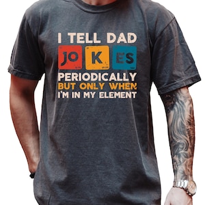 May include: Dark gray t-shirt with the text "I TELL DAD JOKES PERIODICALLY BUT ONLY WHEN I'M IN MY ELEMENT." The text is in white, with the words "JOKES" in a periodic table style. The shirt is worn by a person.