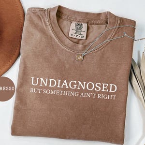 May include: Espresso-colored t-shirt with the words "UNDIAGNOSED BUT SOMETHING AIN'T RIGHT" in white. The shirt has a crew neck and a small tag at the neckline. A silver necklace with a pendant is draped across the shirt.