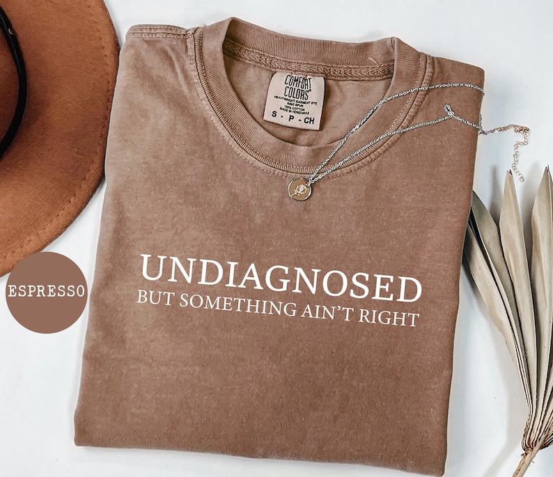 May include: Espresso-colored t-shirt with the words "UNDIAGNOSED BUT SOMETHING AIN'T RIGHT" in white. The shirt has a crew neck and a small tag at the neckline. A silver necklace with a pendant is draped across the shirt.