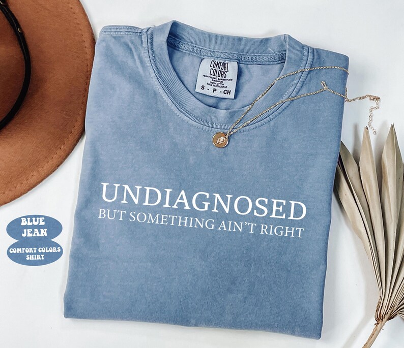 May include: A blue jean Comfort Colors shirt with the text "UNDIAGNOSED BUT SOMETHING AIN'T RIGHT" in white. A gold necklace with a pendant rests on the shirt. The shirt is folded and placed on a white surface.