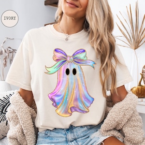 Op de afbeelding: Ivoor kleurig T-shirt met een kleurrijk spookontwerp en een grote strik. Het spook heeft een pastel regenboogkleurenschema met gouden glitteraccenten. Het shirt is een comfortabele, casual stijl.