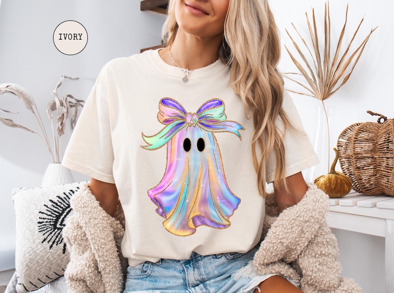 Op de afbeelding: Ivoor kleurig T-shirt met een kleurrijk spookontwerp en een grote strik. Het spook heeft een pastel regenboogkleurenschema met gouden glitteraccenten. Het shirt is een comfortabele, casual stijl.