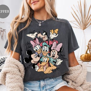 May include: Dark gray t-shirt featuring a graphic of Disney characters including Mickey Mouse, Minnie Mouse, Donald Duck, Daisy Duck, Goofy, and Pluto. The shirt has the text "Daisy Duck", "Minnie Mouse", and "Goofy".