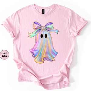 Op de afbeelding: Lichtroze T-shirt met een kleurrijk spookontwerp met een strik. Het spook is versierd met pastelkleuren en gouden glitteraccenten. De tekst "LIGHT PINK" staat in een cirkel aan de linkerkant van het shirt.