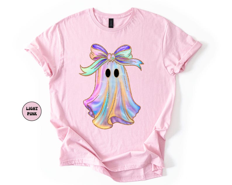 Op de afbeelding: Lichtroze T-shirt met een kleurrijk spookontwerp met een strik. Het spook is versierd met pastelkleuren en gouden glitteraccenten. De tekst "LIGHT PINK" staat in een cirkel aan de linkerkant van het shirt.