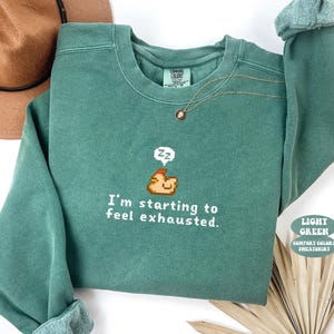 May include: A sage green crewneck sweatshirt featuring a pixel art graphic of a sleeping chicken with a speech bubble containing Zz symbols and the white text I'm starting to feel exhausted. The garment has a ribbed collar and long sleeves.