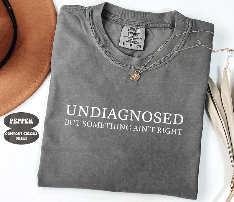 May include: A gray Comfort Colors t-shirt with the text "UNDIAGNOSED BUT SOMETHING AIN'T RIGHT" in white. A gold necklace with a pendant is draped across the shirt. A brown hat is visible in the upper left corner.