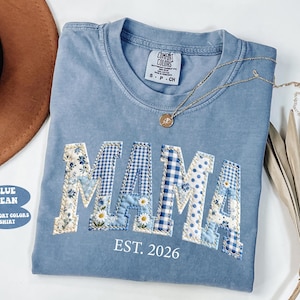 May include: A blue jean Comfort Colors shirt with the word "MAMA" in patchwork letters. The letters are decorated with floral, gingham, and polka dot patterns. The text "EST. 2026" is below the word "MAMA". A gold necklace is draped across the shirt.