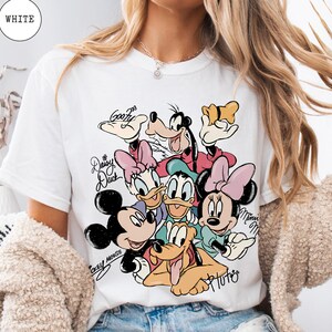 May include: White t-shirt featuring a colorful graphic of Disney characters including Mickey Mouse, Minnie Mouse, Donald Duck, Daisy Duck, Goofy, and Pluto. The shirt has the word "WHITE" in a circle at the top.
