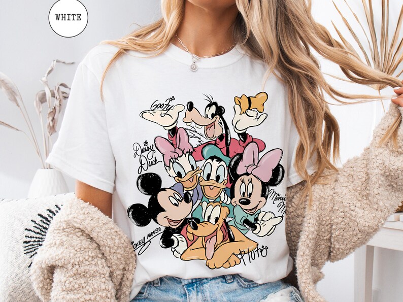 May include: White t-shirt featuring a colorful graphic of Disney characters including Mickey Mouse, Minnie Mouse, Donald Duck, Daisy Duck, Goofy, and Pluto. The shirt has the word "WHITE" in a circle at the top.