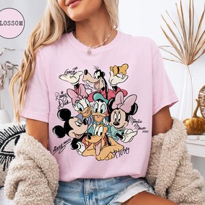 May include: Pink t-shirt featuring a colorful graphic of Disney characters including Mickey Mouse, Minnie Mouse, Donald Duck, Daisy Duck, Goofy, and Pluto. The shirt has the word "Blossom" in a pink circle.