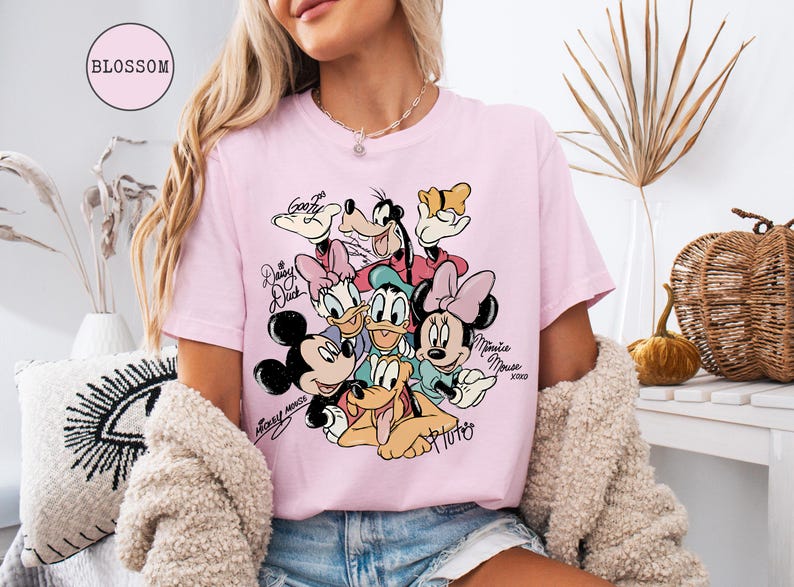 May include: Pink t-shirt featuring a colorful graphic of Disney characters including Mickey Mouse, Minnie Mouse, Donald Duck, Daisy Duck, Goofy, and Pluto. The shirt has the word "Blossom" in a pink circle.