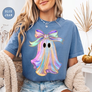 Op de afbeelding: Blauw spijkerbroekkleurig T-shirt met een kleurrijk spookontwerp met een strik. Het spook heeft een glanzende, iriserende uitstraling met zwarte ogen. De tekst "BLUE JEAN" staat in een blauwe cirkel linksboven.