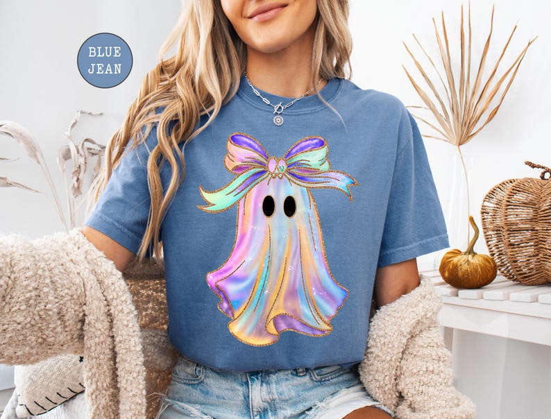 Op de afbeelding: Blauw spijkerbroekkleurig T-shirt met een kleurrijk spookontwerp met een strik. Het spook heeft een glanzende, iriserende uitstraling met zwarte ogen. De tekst "BLUE JEAN" staat in een blauwe cirkel linksboven.