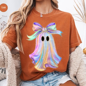 Op de afbeelding: Oranje-bruin T-shirt met een kleurrijk spookontwerp en een strik. De geest is een mix van pastelkleuren met gouden glitteraccenten. Het ontwerp is gecentreerd op het shirt, wat zorgt voor een feestelijke en grillige uitstraling. Het shirt wordt gecombineerd met een spijkerbroek.