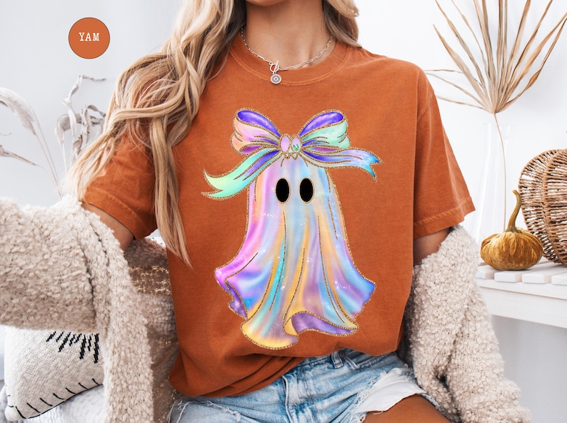 Op de afbeelding: Oranje-bruin T-shirt met een kleurrijk spookontwerp en een strik. De geest is een mix van pastelkleuren met gouden glitteraccenten. Het ontwerp is gecentreerd op het shirt, wat zorgt voor een feestelijke en grillige uitstraling. Het shirt wordt gecombineerd met een spijkerbroek.