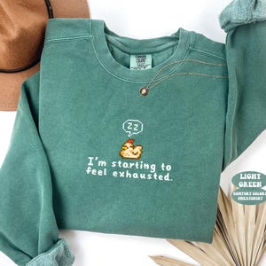 May include: A light green Comfort Colors sweatshirt with the text "I'm starting to feel exhausted." and a pixelated chicken graphic. The sweatshirt has a crew neck and long sleeves. A gold necklace is visible.