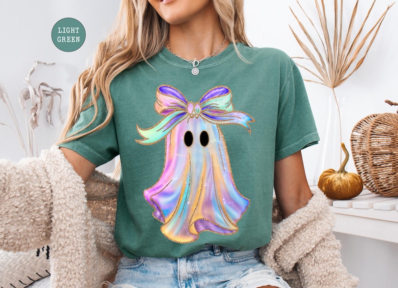 Op de afbeelding: Lichtgroen T-shirt met een kleurrijk spookontwerp en een grote strik. Het spook is afgebeeld in tinten paars, blauw en geel met zwarte ogen. De strik is een mix van paars, groen en geel. Het shirt heeft een comfortabele pasvorm.