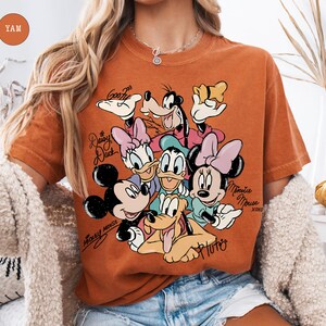 May include: Burnt orange t-shirt featuring a graphic of Mickey Mouse, Minnie Mouse, Donald Duck, Daisy Duck, Goofy, and Pluto. The shirt has the text "Mickey Mouse" and "Minnie Mouse" in cursive. The shirt is a crew neck and has short sleeves.