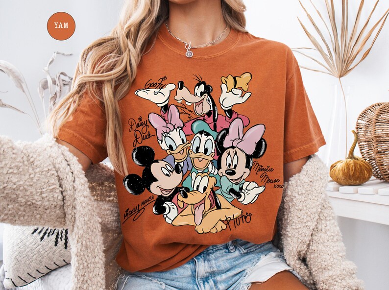 May include: Burnt orange t-shirt featuring a graphic of Mickey Mouse, Minnie Mouse, Donald Duck, Daisy Duck, Goofy, and Pluto. The shirt has the text "Mickey Mouse" and "Minnie Mouse" in cursive. The shirt is a crew neck and has short sleeves.
