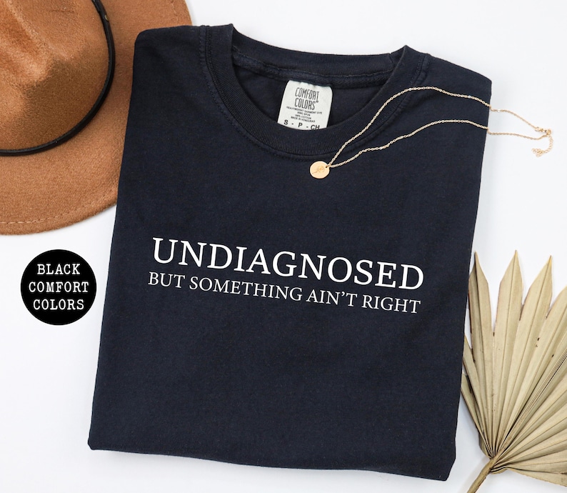 May include: Black crew neck sweatshirt with the text "UNDIAGNOSED BUT SOMETHING AIN'T RIGHT" in white. A gold necklace with a small pendant rests on the shirt. A brown hat and dried palm leaves are also in the image.