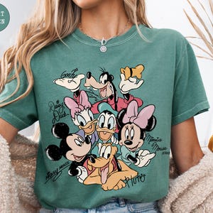 May include: Light green t-shirt featuring a colorful graphic of Disney characters including Mickey Mouse, Minnie Mouse, Donald Duck, Daisy Duck, Goofy, and Pluto. The characters' names are written in cursive. The shirt is a comfortable, casual style.