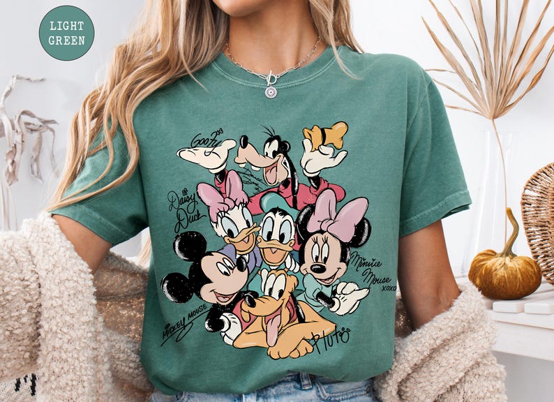 May include: Light green t-shirt featuring a colorful graphic of Disney characters including Mickey Mouse, Minnie Mouse, Donald Duck, Daisy Duck, Goofy, and Pluto. The characters' names are written in cursive. The shirt is a comfortable, casual style.
