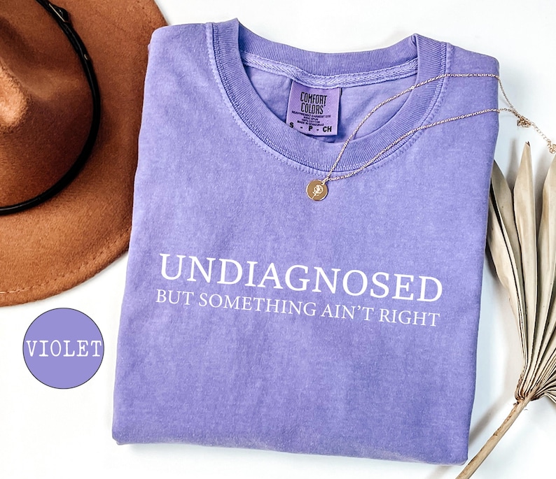 May include: A violet Comfort Colors t-shirt with the text "UNDIAGNOSED BUT SOMETHING AIN'T RIGHT" in white. A gold necklace and a brown hat are also in the image. The shirt is a size small.
