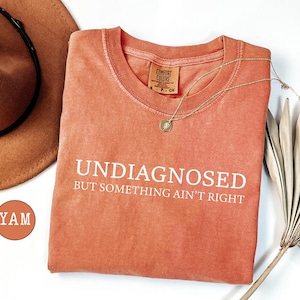 May include: Coral-colored t-shirt with the text "UNDIAGNOSED BUT SOMETHING AIN'T RIGHT" in white. A gold necklace and a brown hat are also visible. The shirt is made of a soft material and has a round neckline.