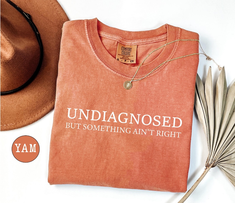 May include: Coral-colored t-shirt with the text "UNDIAGNOSED BUT SOMETHING AIN'T RIGHT" in white. A gold necklace and a brown hat are also visible. The shirt is made of a soft material and has a round neckline.