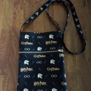 May include: Black crossbody bag with a zip closure. The bag is made of fabric with a repeating pattern of Harry Potter and Gryffindor logos, lightning bolts, and glasses.