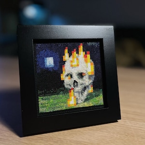 May include: A framed cross-stitch artwork depicting a white skull with flames on top, set against a dark blue background with a white square in the top left corner. The skull is surrounded by green grass.
