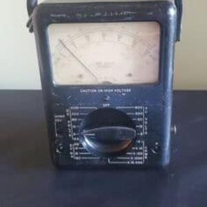 Puede incluir: Un multímetro analógico vintage negro con un dial redondo y una perilla. El dial tiene una aguja y marcas para voltaje, corriente y resistencia. El multímetro tiene una caja de plástico negro con un asa.