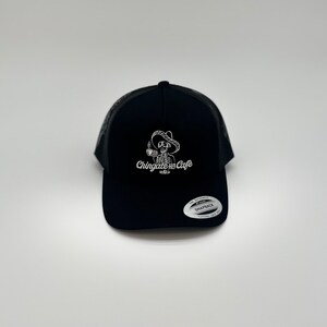 Può includere: Cappellino da camionista nero con un designo ricamato bianco di uno scheletro che indossa un sombrero e tiene una tazza di caffè. Il testo "Chingate un Cafe" è ricamato sotto lo scheletro.