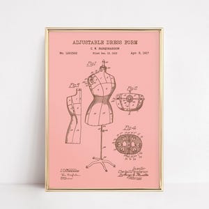May include: A framed print featuring a vintage illustration of an adjustable dress form, with detailed diagrams and text. The artwork is in sepia tones against a soft pink background, encased in a gold frame. The print includes the text "ADJUSTABLE DRESS FORM".