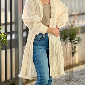 May include: A long-sleeved, off-white, lightweight cotton shirt with a button-up front and a ruffled hem. The shirt is worn over a brown tank top and blue jeans.