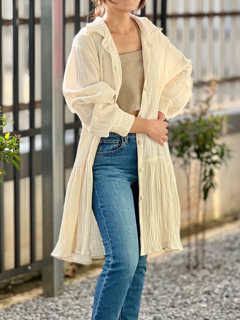 May include: A long-sleeved, off-white, lightweight cotton shirt with a button-up front and a ruffled hem. The shirt is worn over a brown tank top and blue jeans.