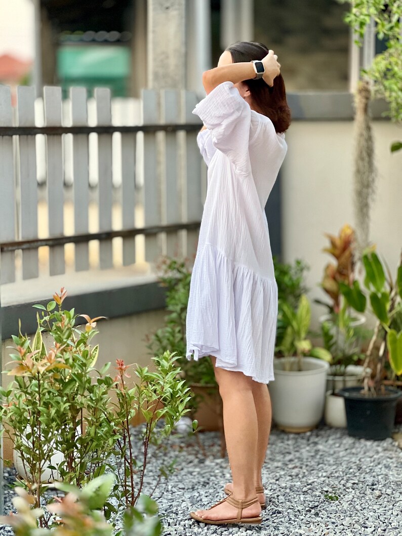 May include: A woman wearing a white, flowing dress with a ruffled hem. She is standing in a garden setting with green plants and gravel.