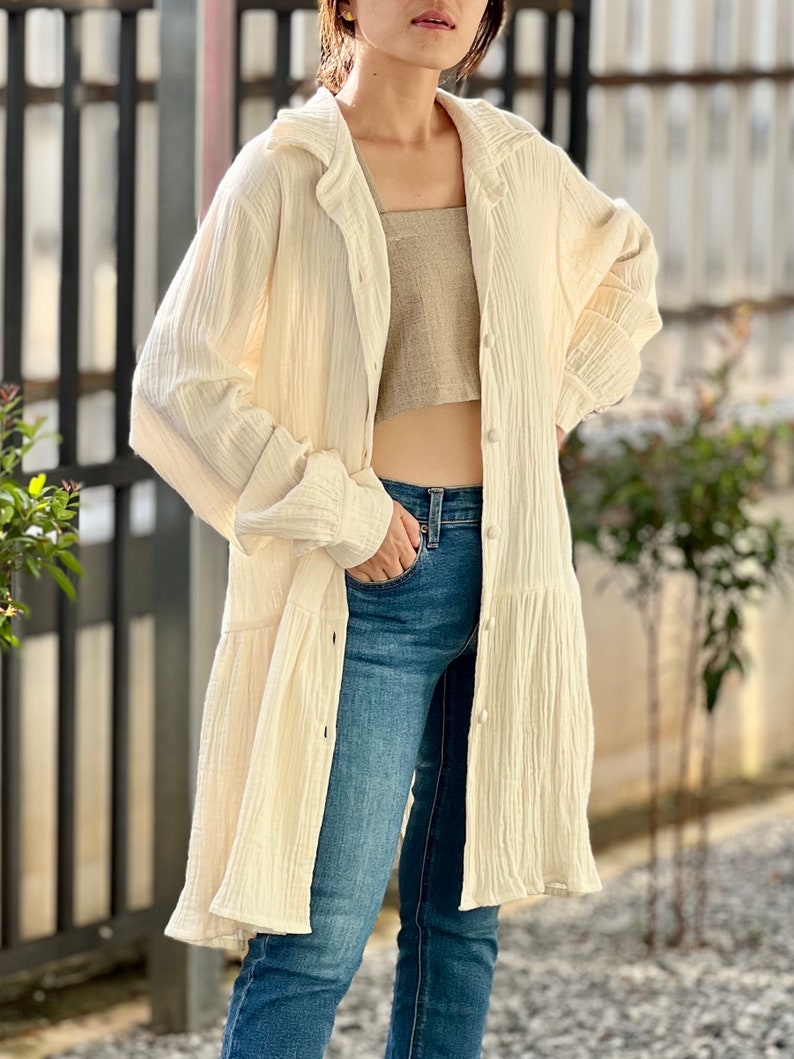 May include: A white, long-sleeved, button-down shirt with a ruffled hem. The shirt is made of a lightweight, textured fabric and has a relaxed fit.