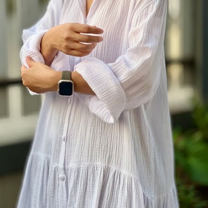 May include: A white, long-sleeved, button-down shirt dress with a ruffled hem. The dress is made of a lightweight, textured fabric.