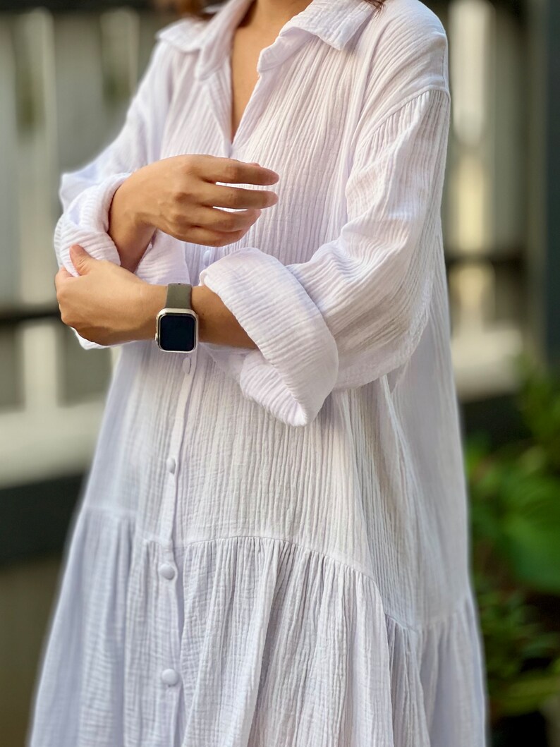 May include: A white, long-sleeved, button-down shirt dress with a ruffled hem. The dress is made of a lightweight, textured fabric.
