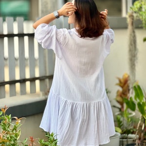 May include: A white, loose-fitting dress with a ruffled hem. The dress is made of a lightweight, breathable fabric and has a relaxed fit. The model is wearing the dress with a watch on her wrist.