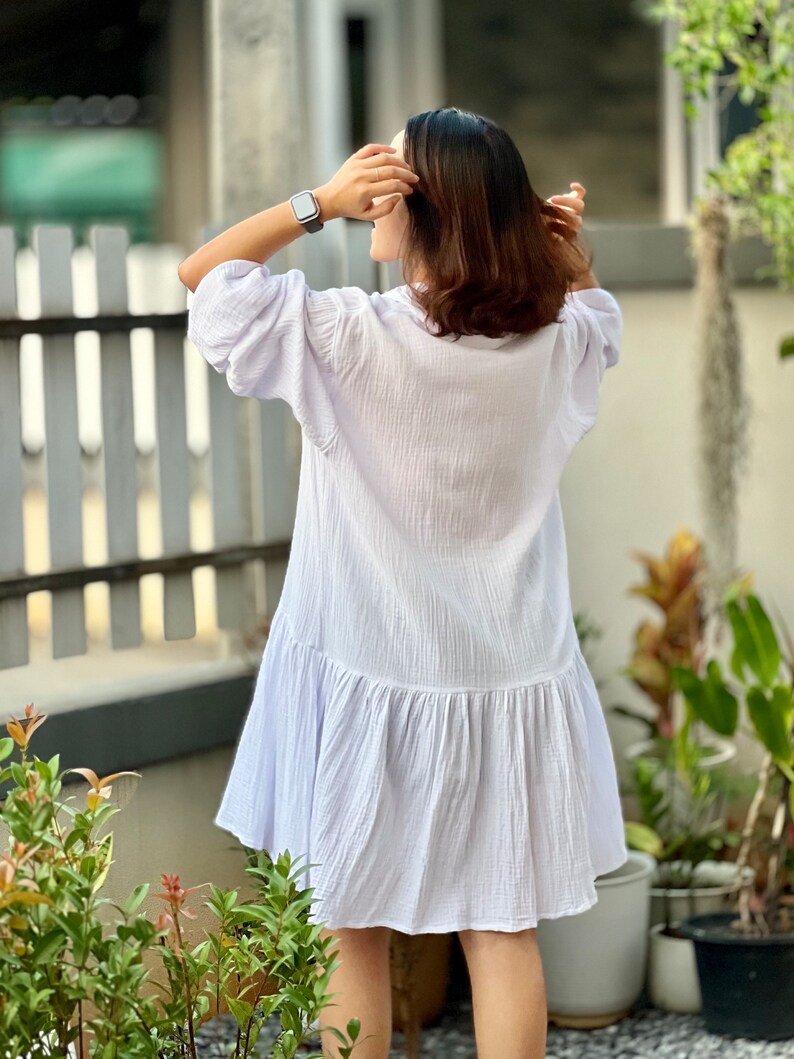 May include: A white, loose-fitting dress with a ruffled hem. The dress is made of a lightweight, breathable fabric and has a relaxed fit. The model is wearing the dress with a watch on her wrist.
