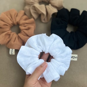 Puede incluir: Cuatro scrunchies en diferentes colores: beige, marrón, negro y blanco. El scrunchie blanco está hecho de un tejido suave y texturizado y está siendo sostenido por una mano.